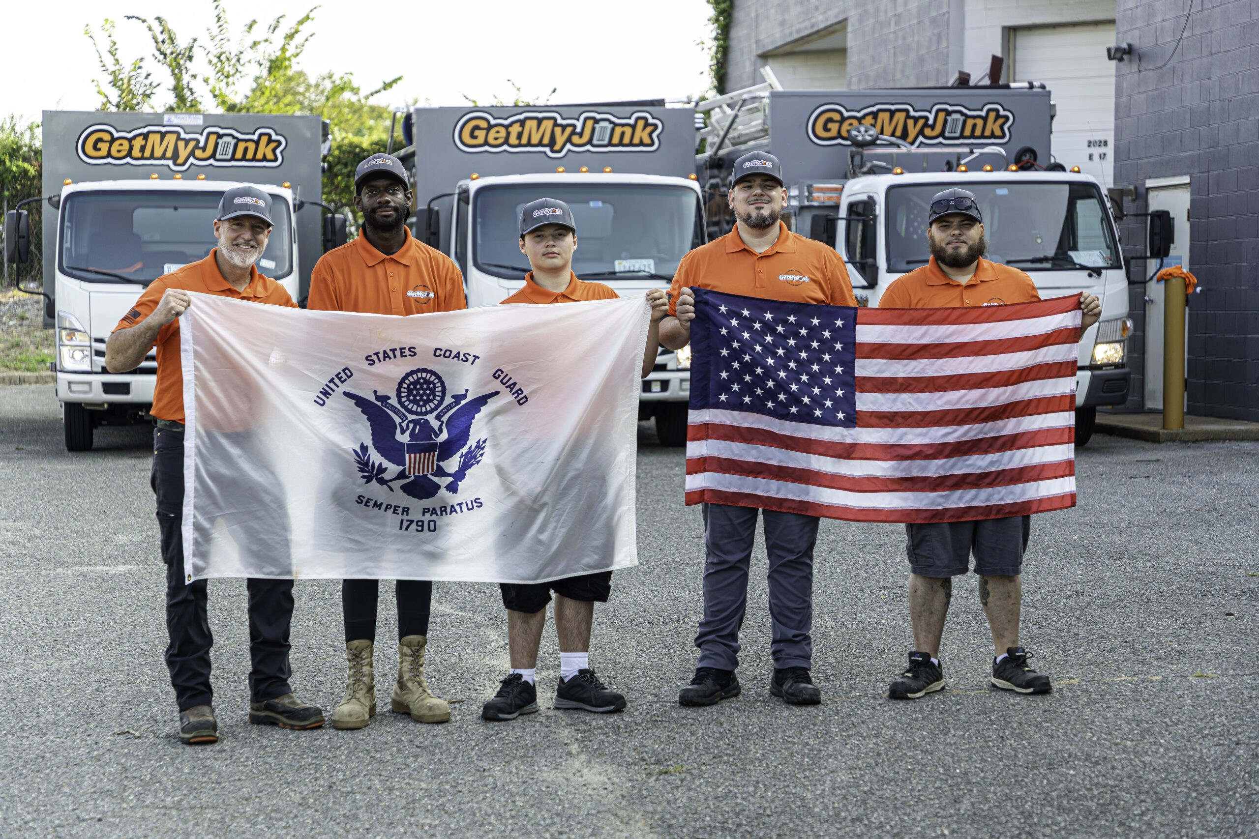 The Get My Junk junk removal team holds up an American flag and United States Coast Guard flag with junk removal trucks in the background.